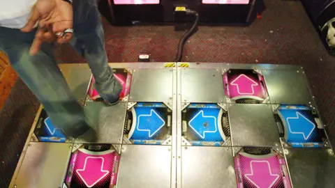 Getty Images A metal Dance Dance Revolution platform showing blue and pink arrows pointing up right down and left. Someone is standing on the platform tapping the up arrow with their right foot.